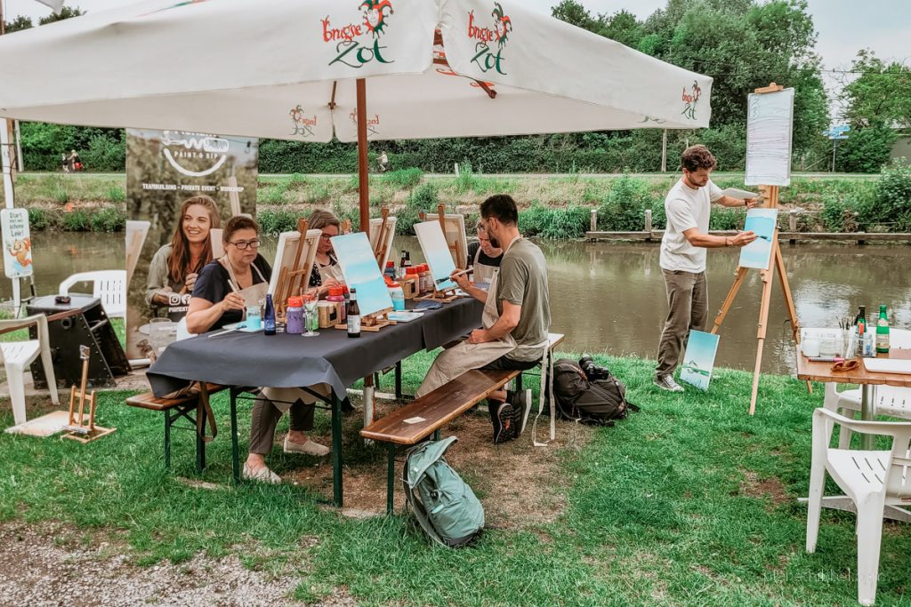 Menschen, die auf Bierbänken unter einem Schirm sitzen und an Staffeleien malen. Im Hintergrund der Fluss Leie und ein Mal-Lehrer, der an einer Staffelei Instruktionen gibt. 