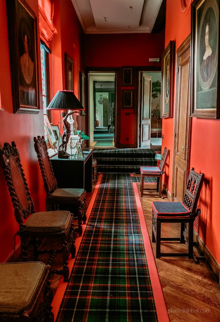 Flur mit roten Wänden, karrierten Teppichen, Bildern und Stühlen im Schloss Ooidonk. 