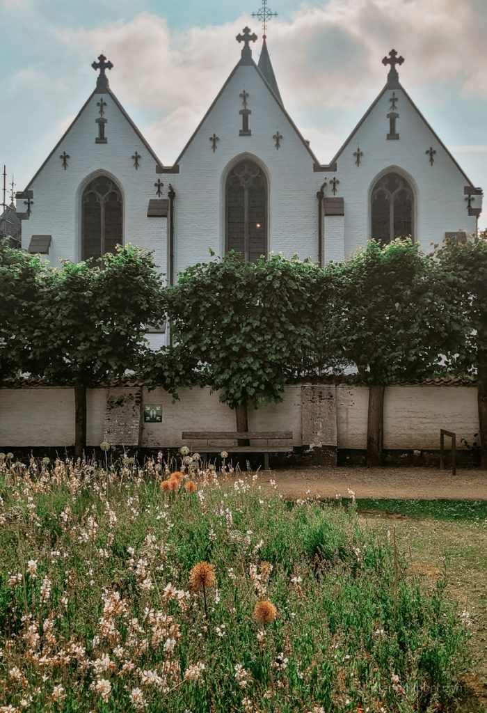 Weiße Kirche mit drei Giebeln in Sint-Martens-Laatem. Im Vordergrund Bäume und Blumenwiese. 